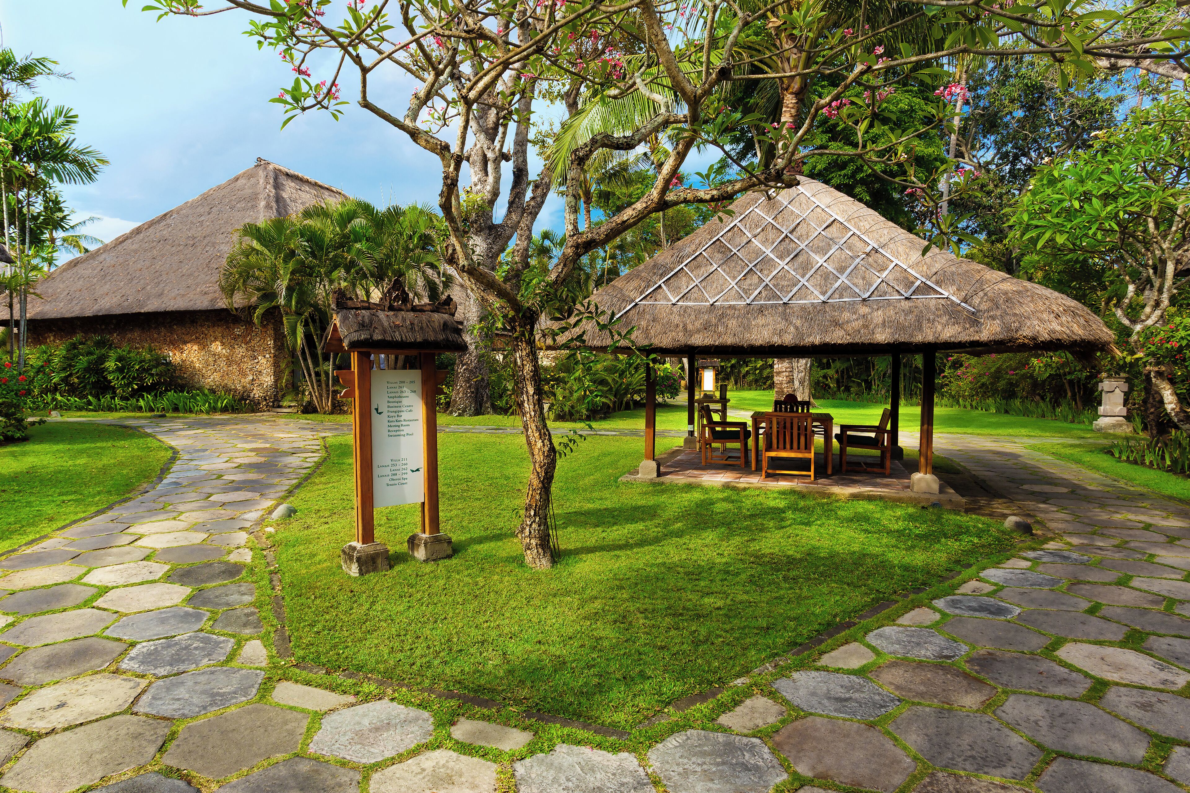 A stone pathway leading to a gazebo with a thatched roof, surrounded by lush greenery and a signpost with information.