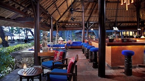 An outdoor seating area with blue cushioned chairs and tables, a thatched roof, and a bar area with stools. - The Oberoi, Bali (Seminyak)