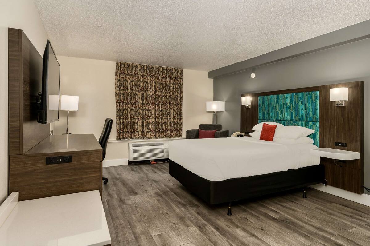 A modern hotel room with a large bed, a desk with a flat-screen TV, a chair, a desk lamp, and a wall-mounted heater.