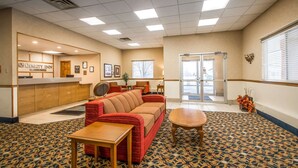 A hotel lobby with a reception desk, a red sofa, a wooden coffee table, patterned carpet, and a large window with blinds.