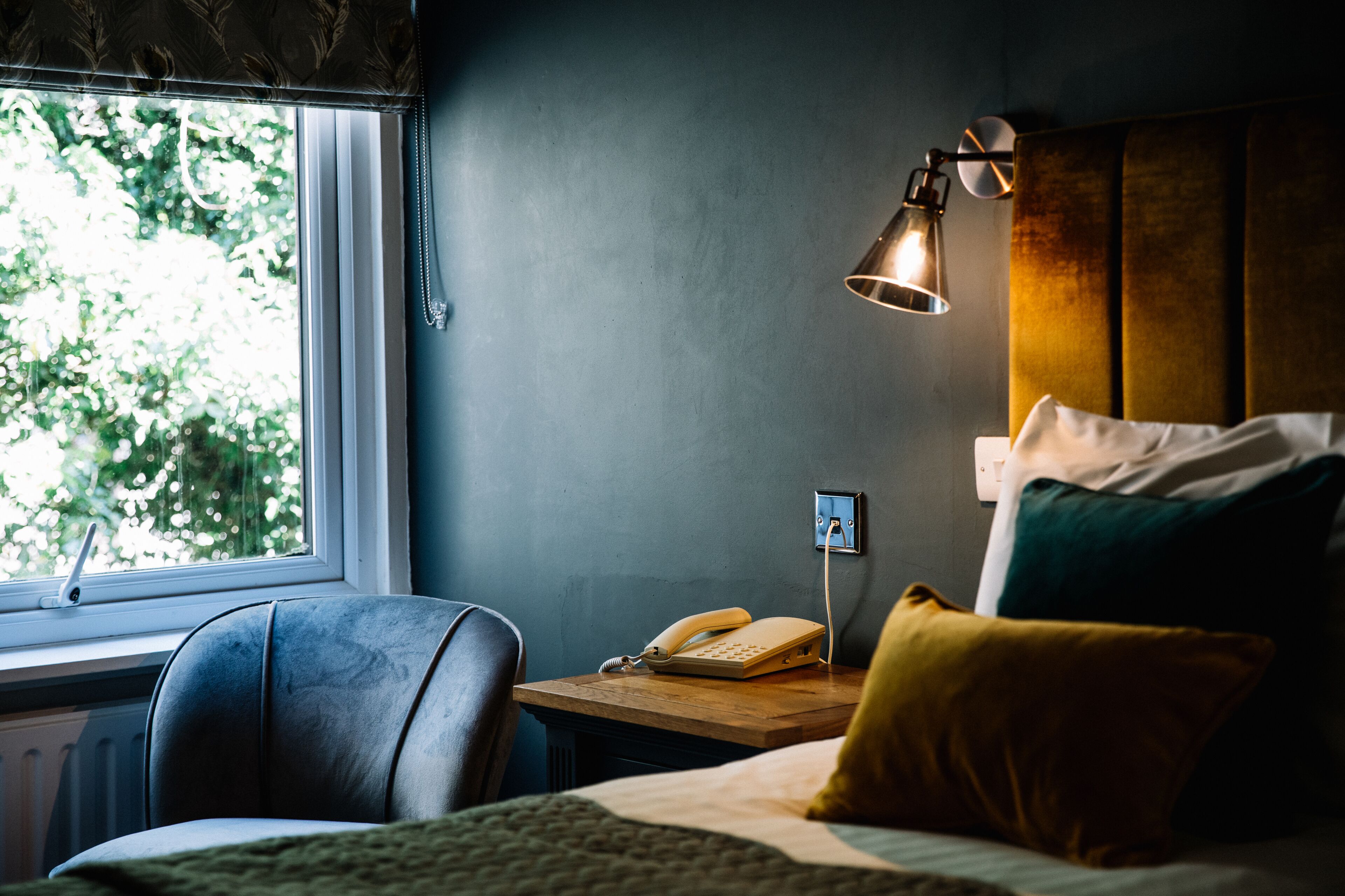 A hotel room with a bed, a desk with a telephone, a lamp, and a window with a view of trees.