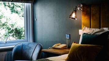 Una habitación de hotel con una cama, un escritorio con teléfono, una lámpara y una ventana con vistas a árboles.