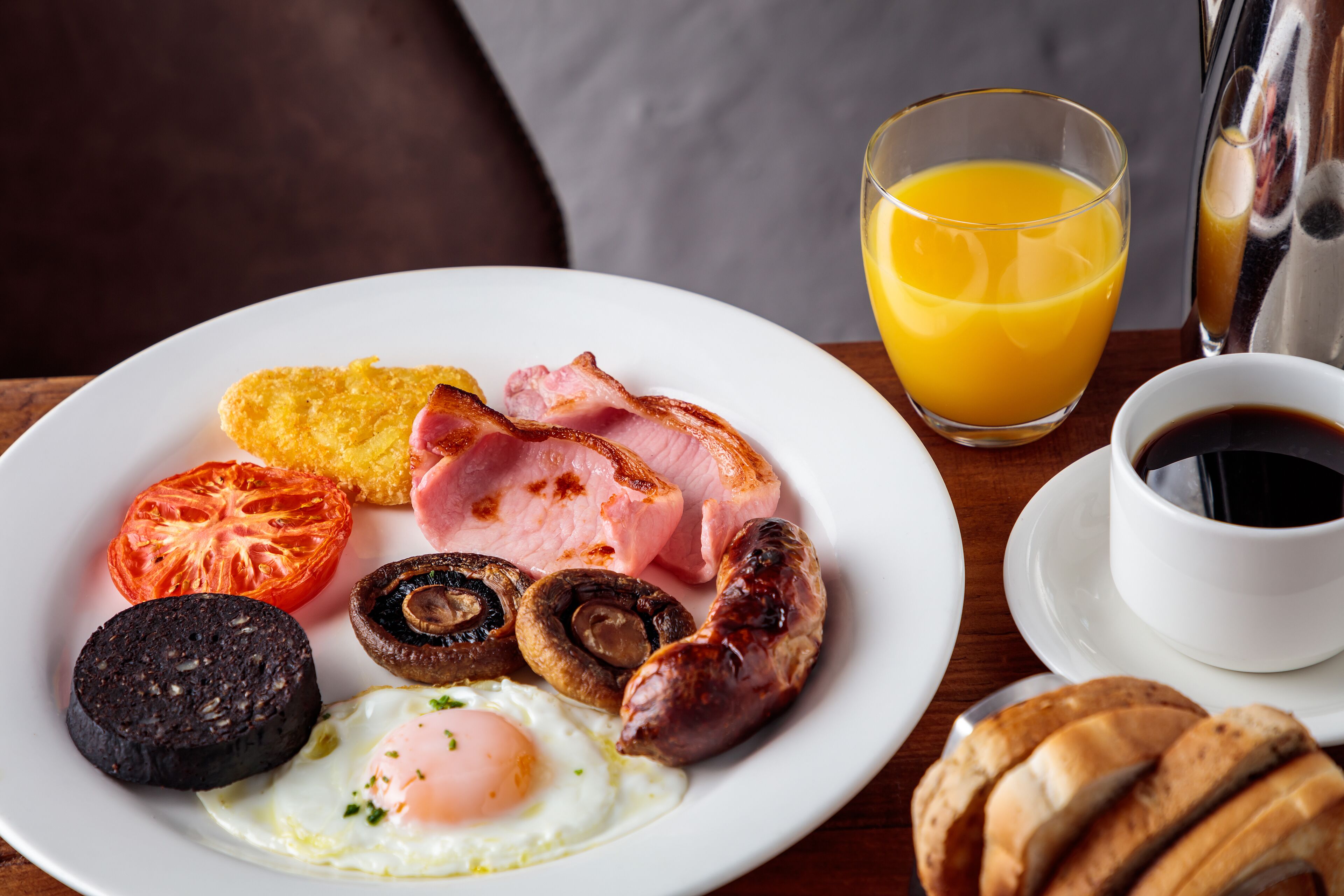 Un piatto per la colazione con uova, pancetta, funghi, salsiccia e pane tostato, insieme a un bicchiere di succo d'arancia e una tazza di caffè.
