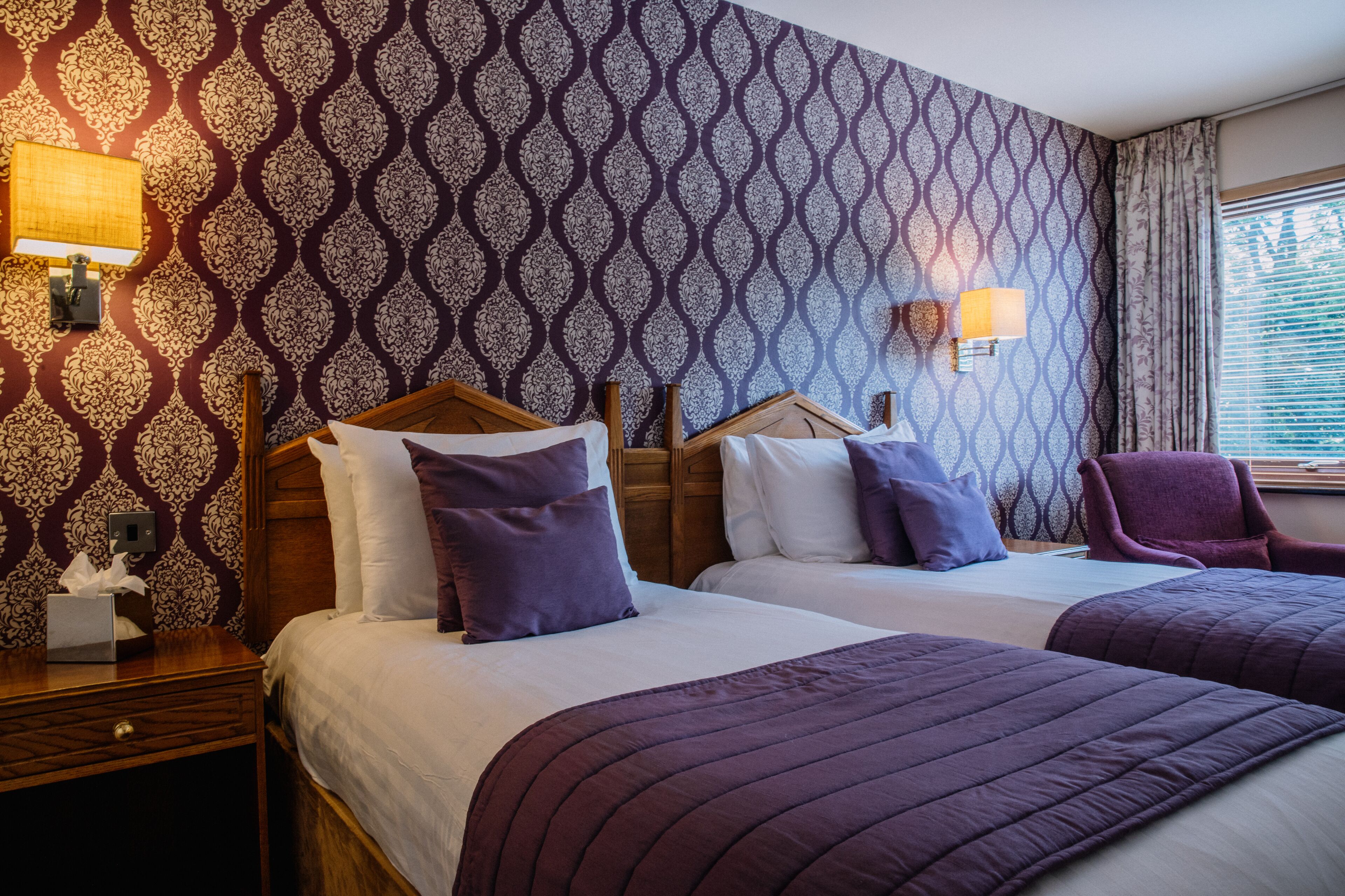 A hotel room with two beds, patterned wallpaper, bedside lamps, a chair, and a nightstand with a tissue box.