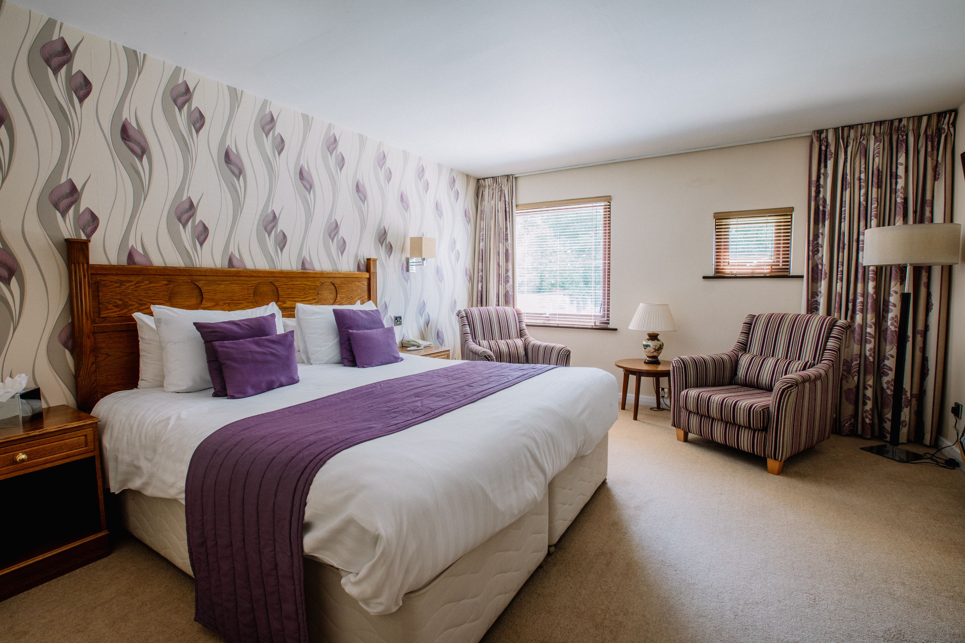 A hotel room with a large bed, two armchairs, a nightstand, and a window with blinds.