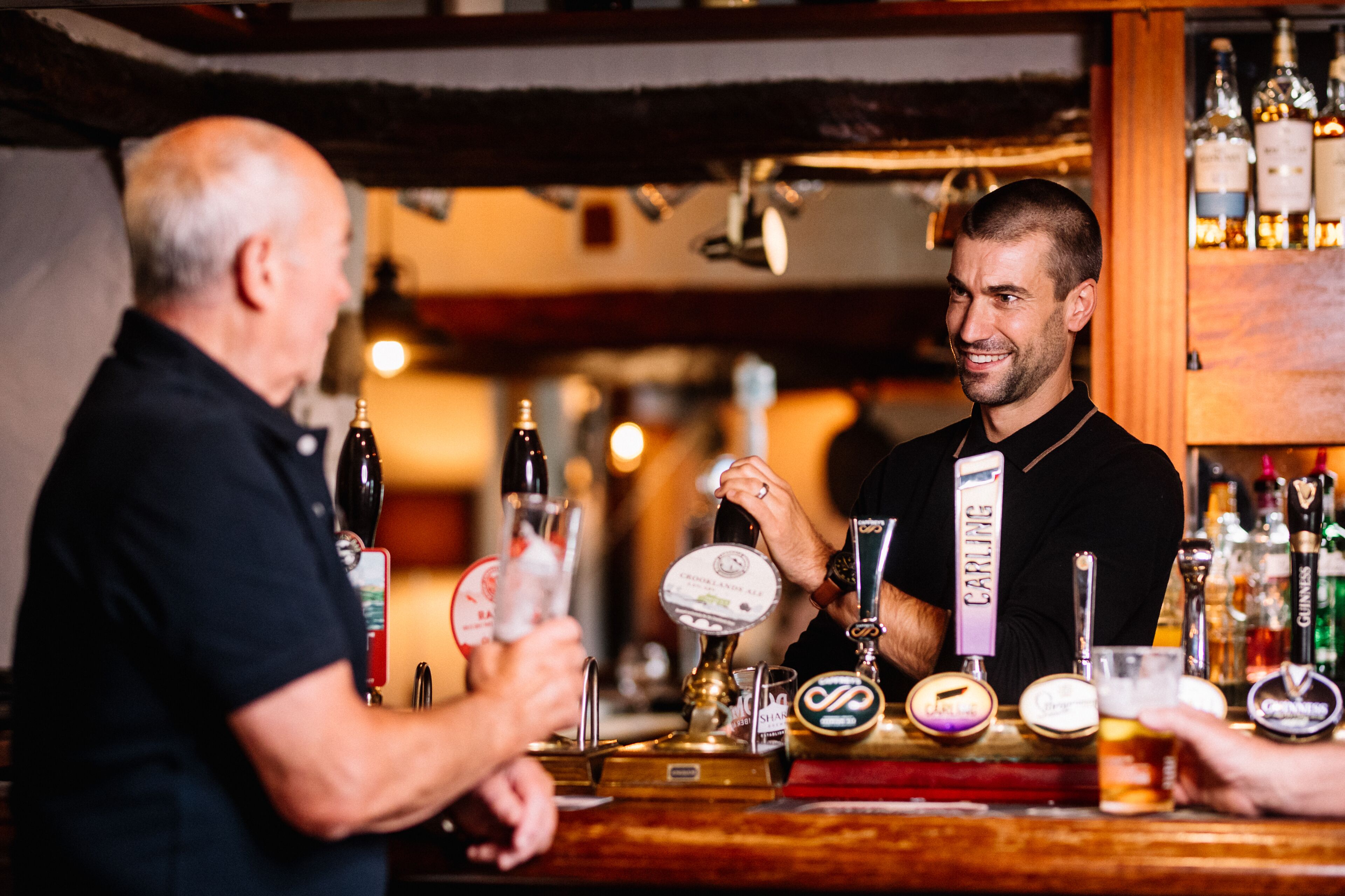 Un bar con un cantinero atendiendo a un cliente. Hay varias botellas y un grifo con la etiqueta "CARLING" en la barra.