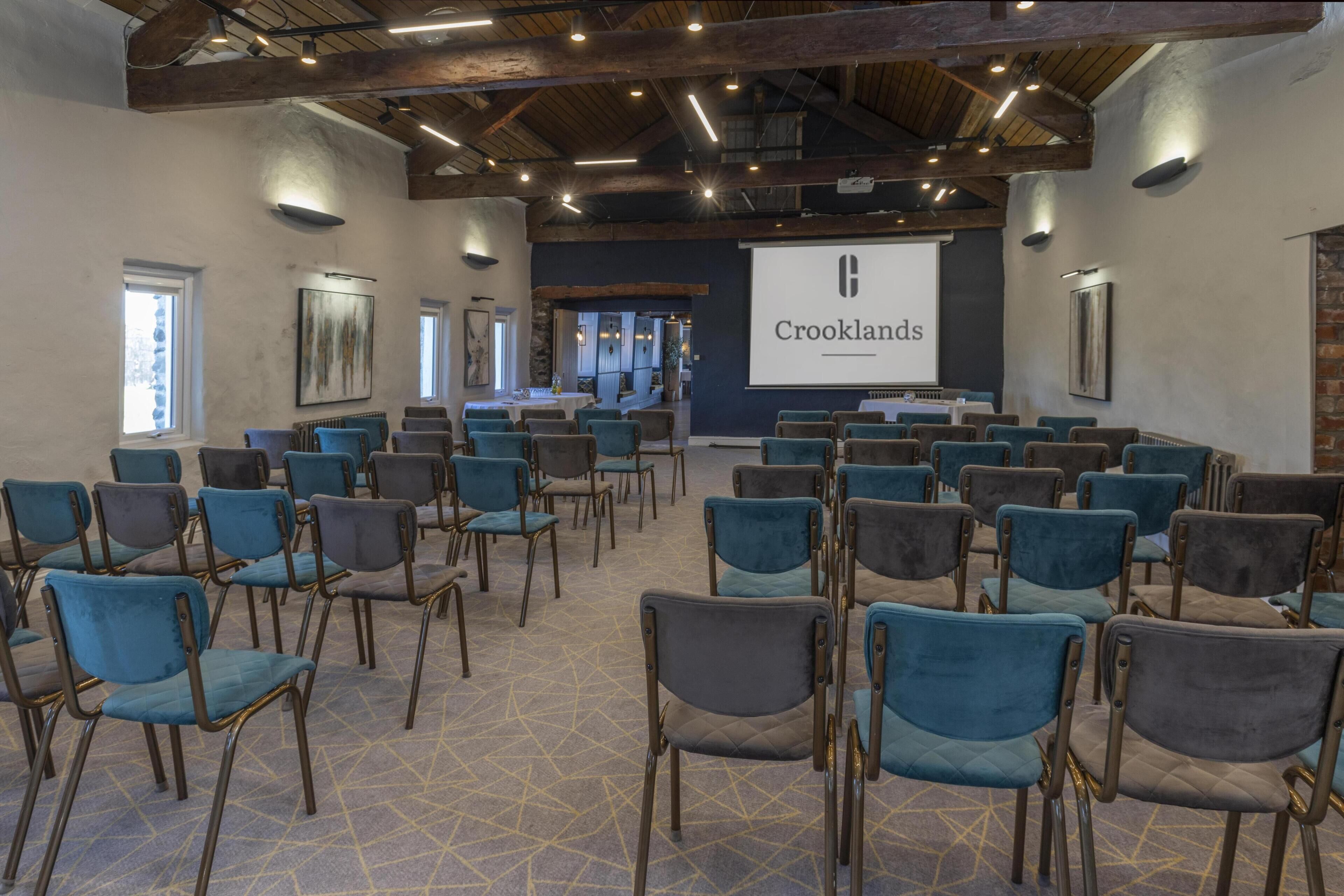 A conference room with rows of chairs, a projector screen displaying 'Crooklands,' and a stone wall.