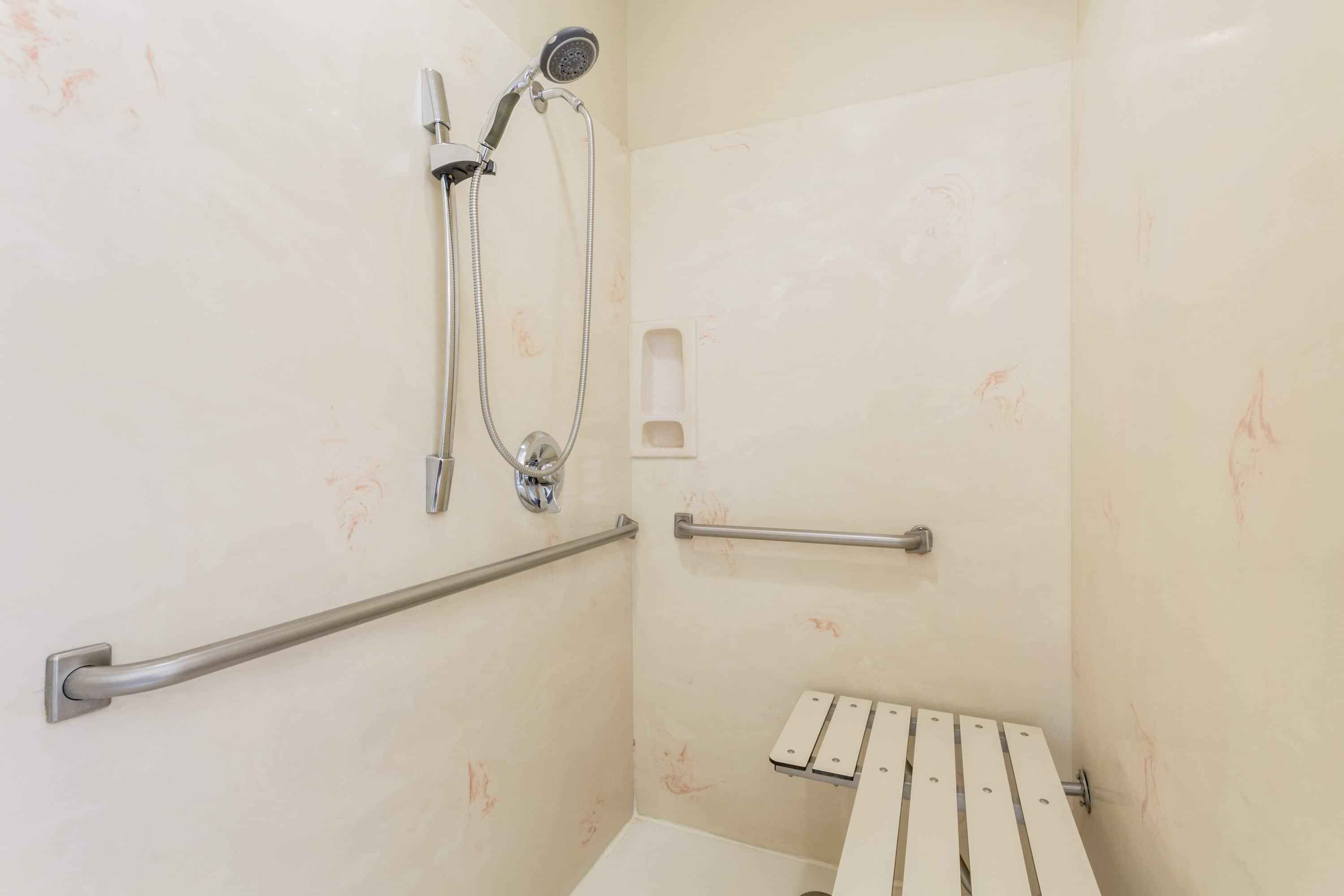 A shower area with a handheld showerhead, a handheld shower attachment, a grab bar, and a white bench.