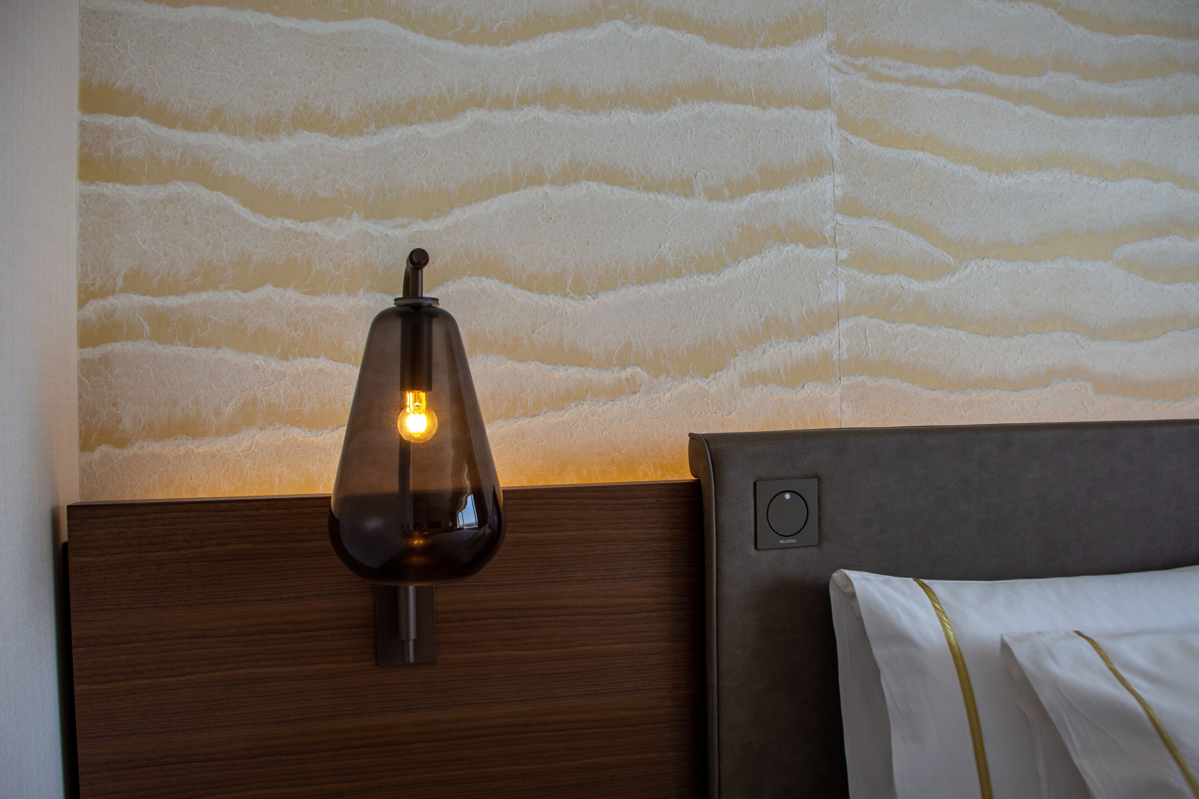 A wall-mounted lamp with a visible light bulb, a wooden headboard, and a textured wall.