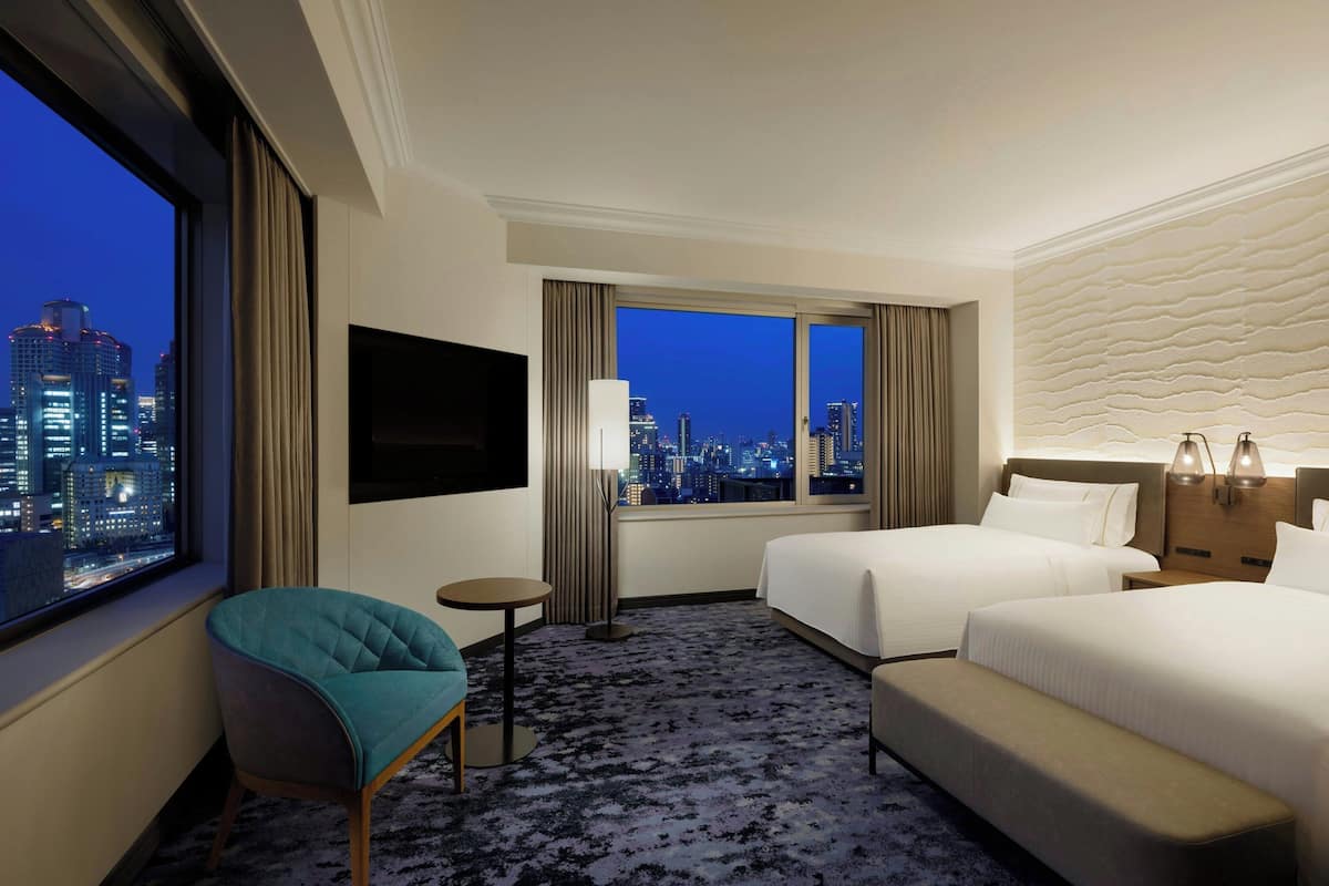 A hotel room with two beds, a TV, a chair, and a view of the city at night.