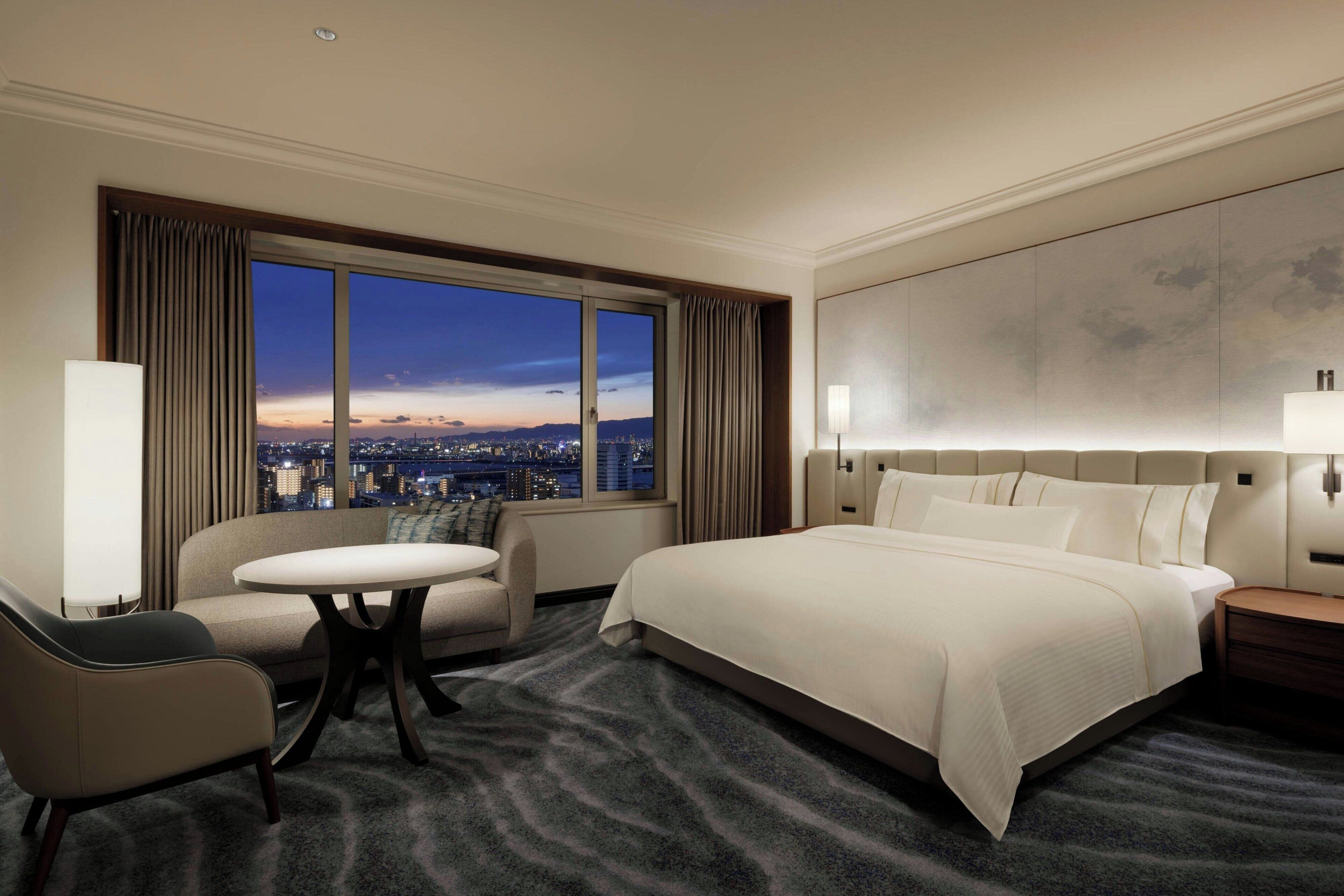 A hotel room with a large bed, a sofa, a round table, and a view of the city at dusk.