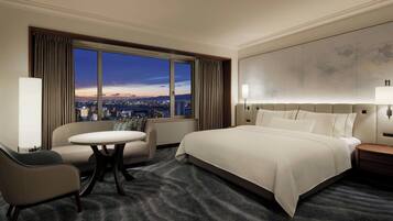 A hotel room with a large bed, a sofa, a round table, and a view of the city at dusk.