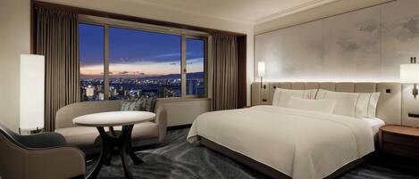 A hotel room with a large bed, a sofa, a round table, and a view of the city at dusk.