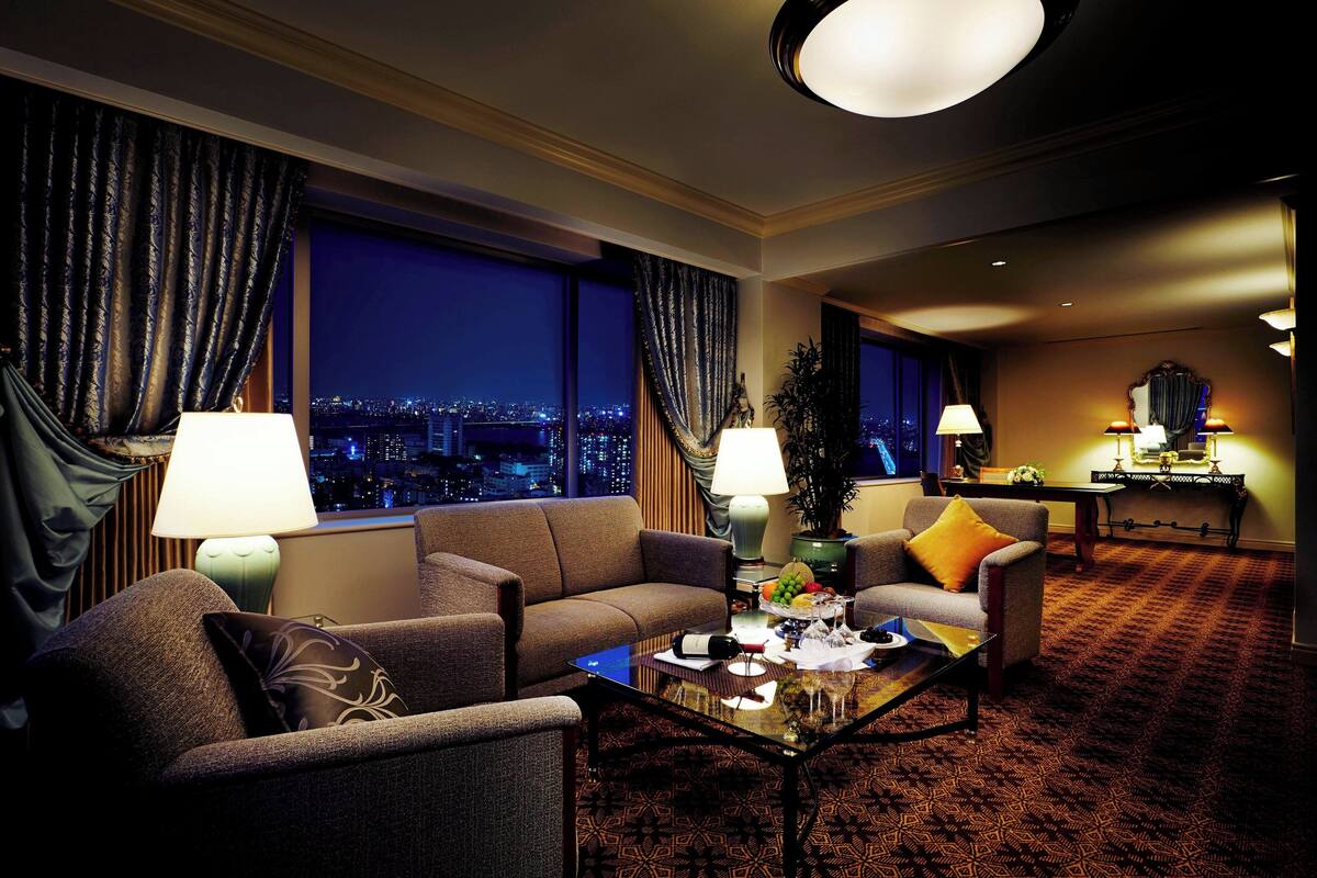 A hotel room with a city view, two sofas, a coffee table, and a dining area with a lamp and table.