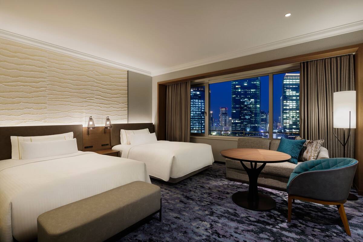 A hotel room with two beds, a sofa, a round table, and a view of a cityscape at night.