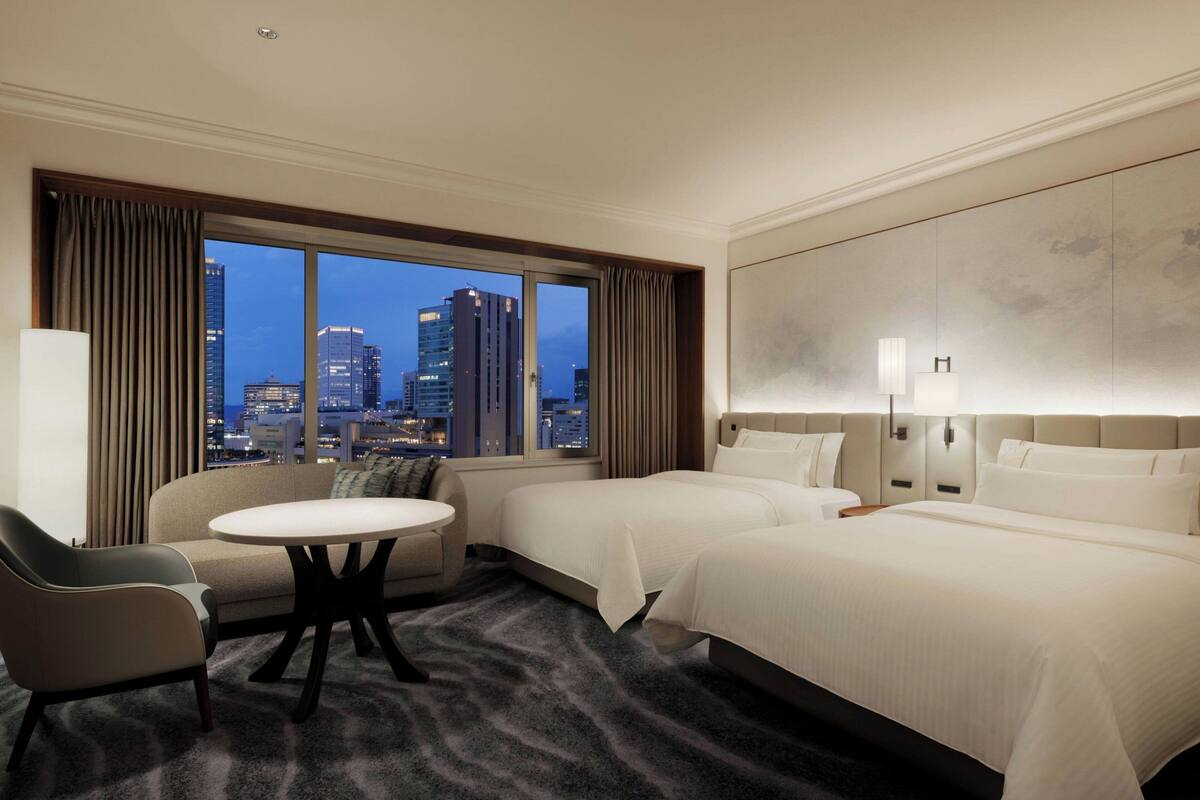 A hotel room with two beds, a sofa, a small table, and a view of the cityscape at dusk.