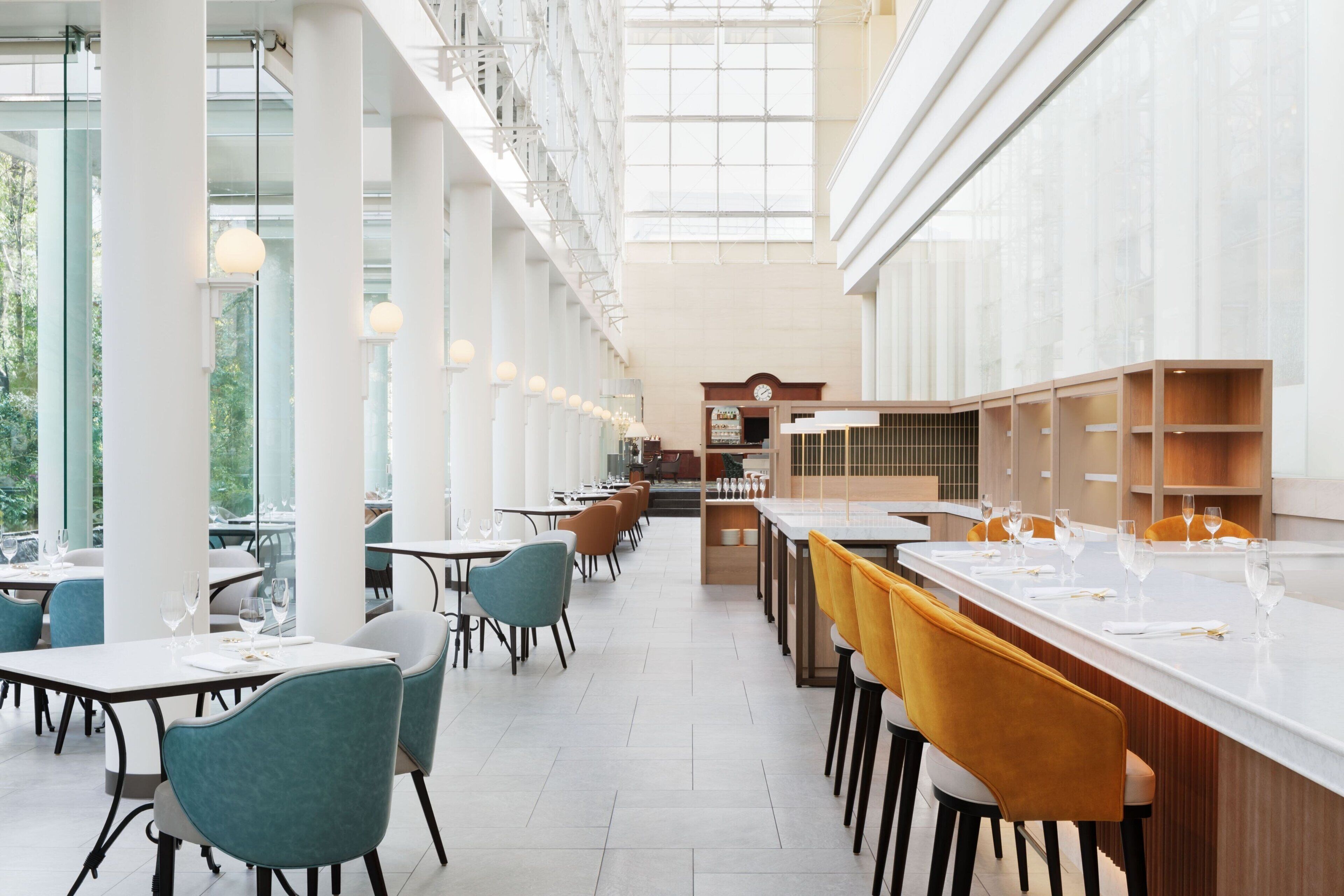 Un restaurante espacioso con techos altos y ventanas amplias que dejan entrar luz natural. Las mesas están ordenadas con sillas, y el interior presenta un diseño moderno con detalles blancos y de madera.