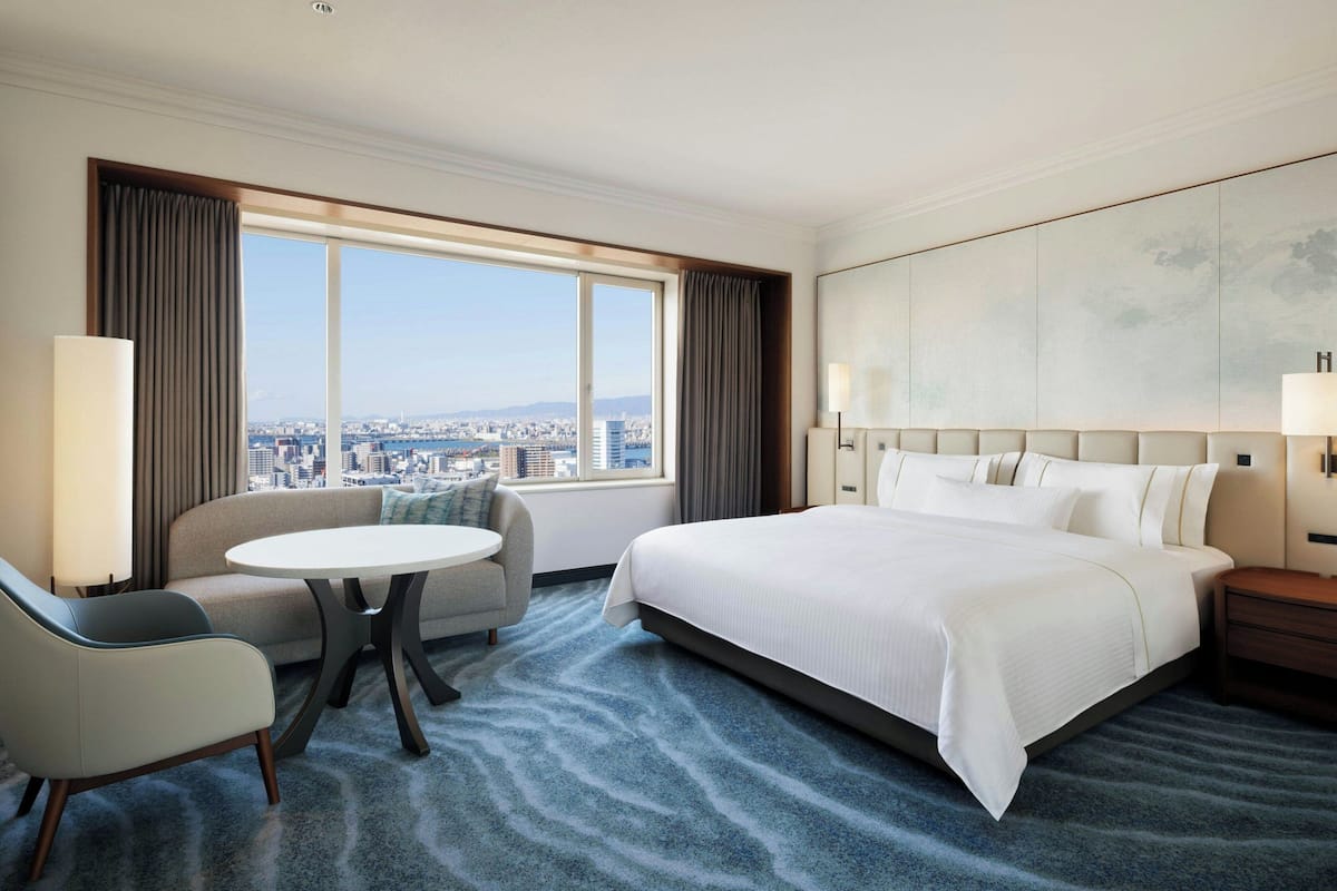 A hotel room with a large bed, a sofa, a small table, and a view of a cityscape through a large window.