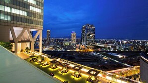 Un paisaje urbano nocturno con un edificio moderno que tiene una terraza en la azotea con zonas de estar y plantas en macetas.