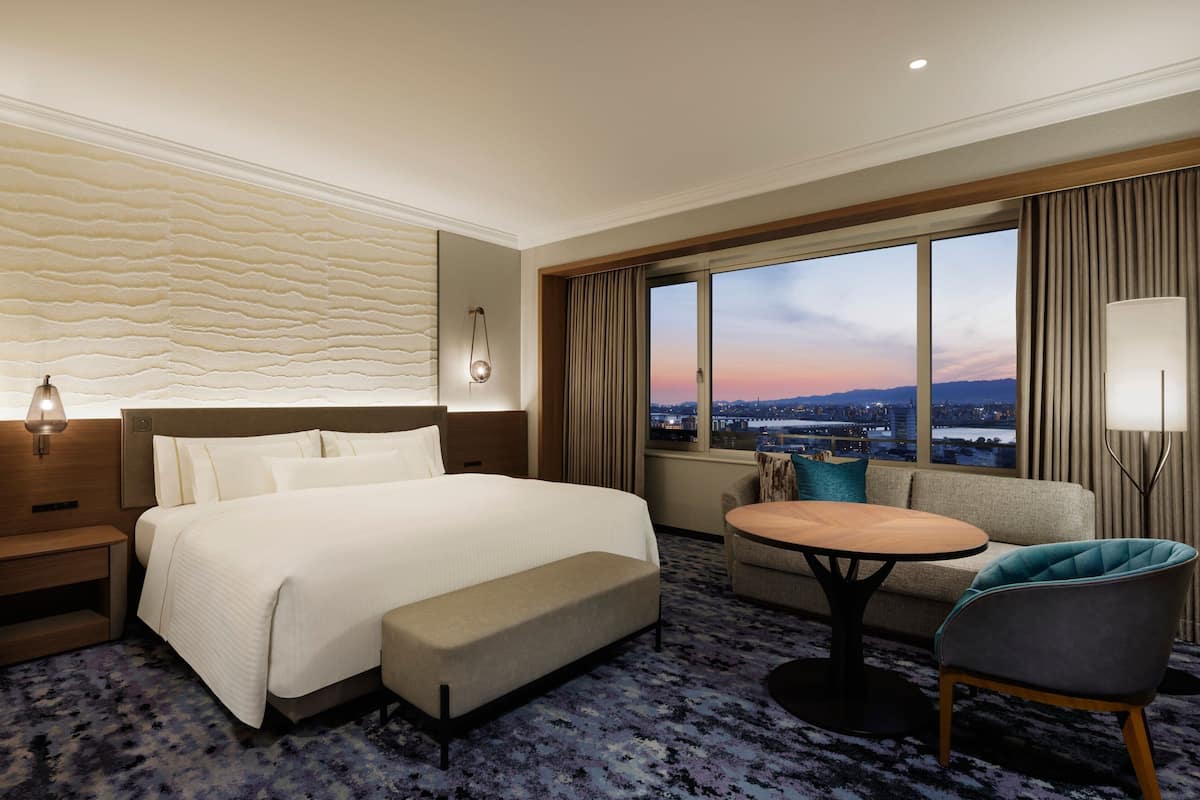 A hotel room with a large bed, a sofa, a small table, and a view of the city at sunset.