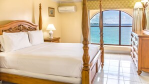 A four-poster bed with white linens, a nightstand with a lamp, a dresser with drawers, and a view of the sea through a large window. - Barbados Beach Club Resort - All Inclusive (Maxwell)