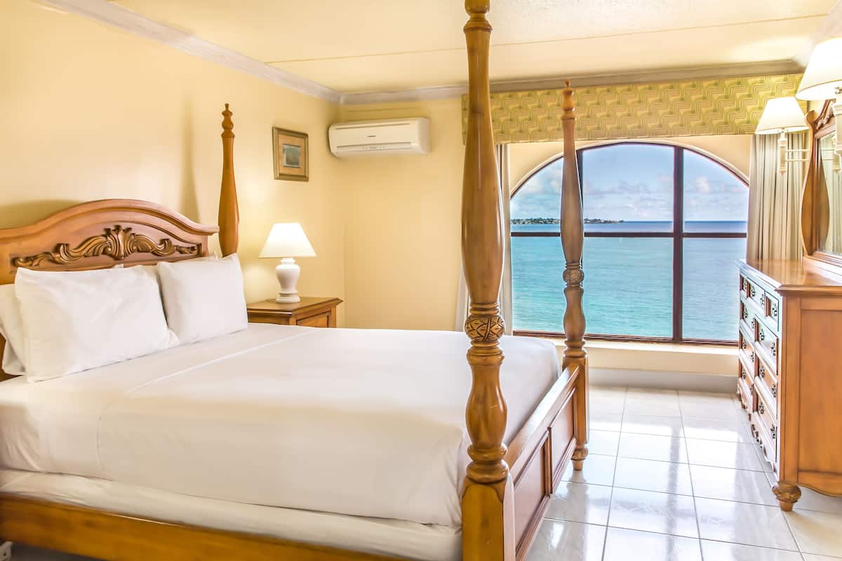 A four-poster bed with white linens, a nightstand with a lamp, a dresser with drawers, and a view of the sea through a large window.