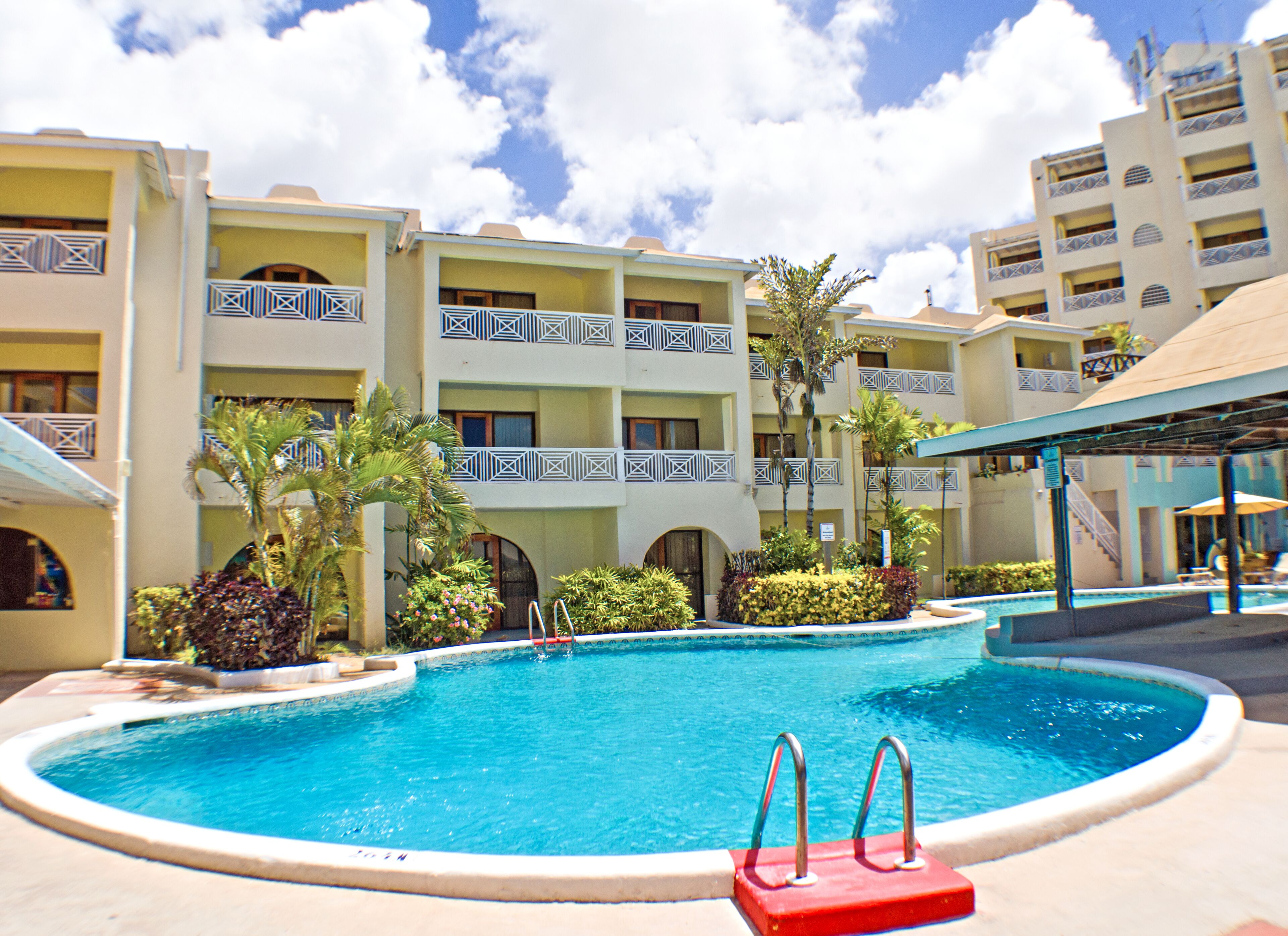 Zona de piscina de hotel con una piscina azul cristalina, rodeada de palmeras y un edificio de varios pisos con balcones.
