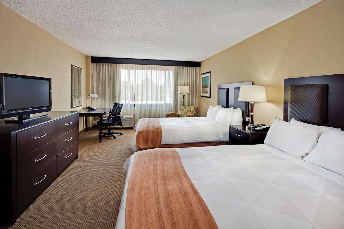 A hotel room with two beds, a desk with a TV, a chair, a lamp, and a window with curtains.