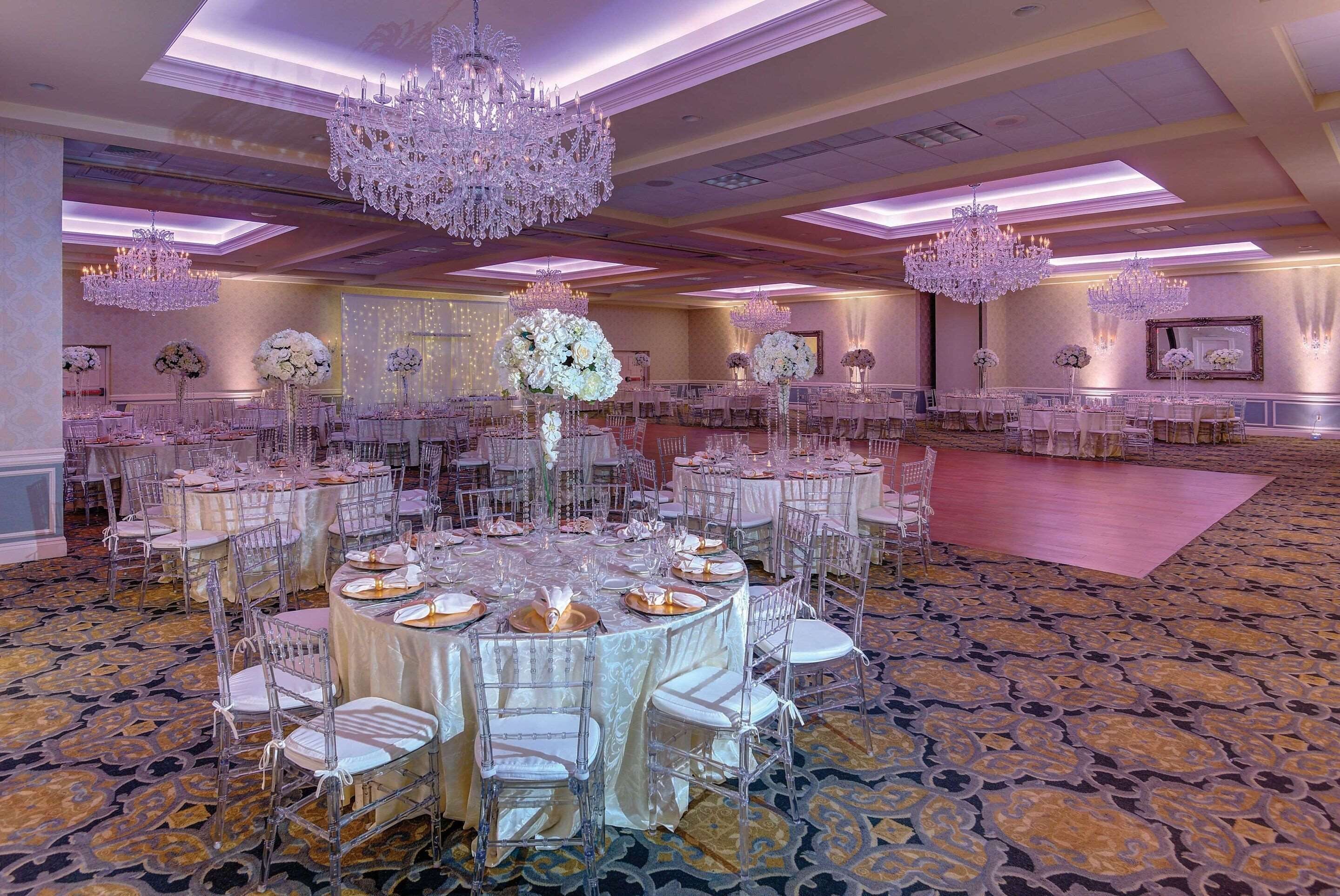 A banquet hall with round tables, clear chairs, and chandeliers.