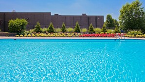 A clear blue swimming pool with a metal ladder, surrounded by a variety of colorful flowers and greenery, and a large building in the background.