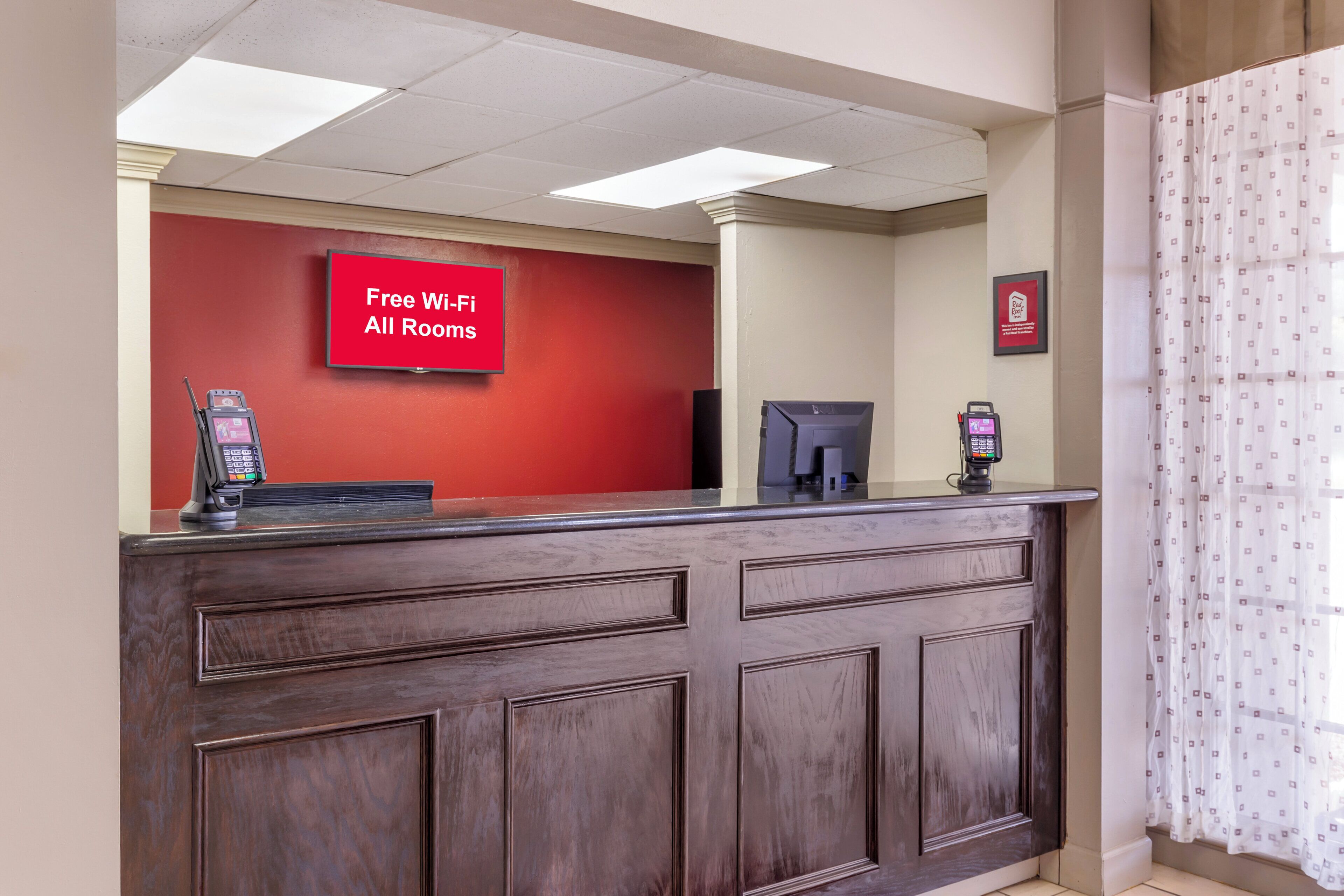 A reception desk with a red wall, a digital screen displaying 'Free Wi-Fi All Rooms,' and a framed sign above it.