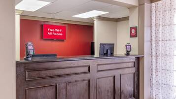 A reception desk with a red wall, a digital screen displaying 'Free Wi-Fi All Rooms,' and a framed sign above it.