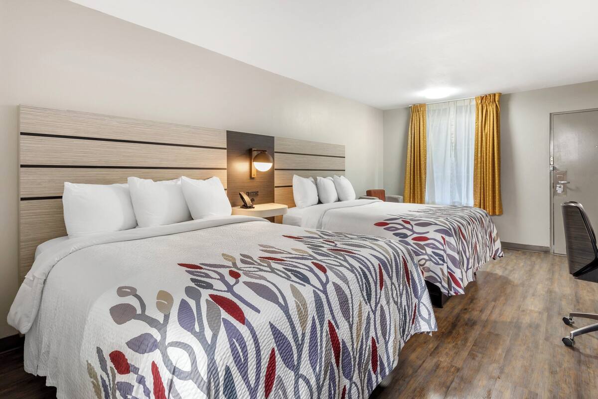 A hotel room with two beds, a nightstand, a lamp, a chair, and a door.