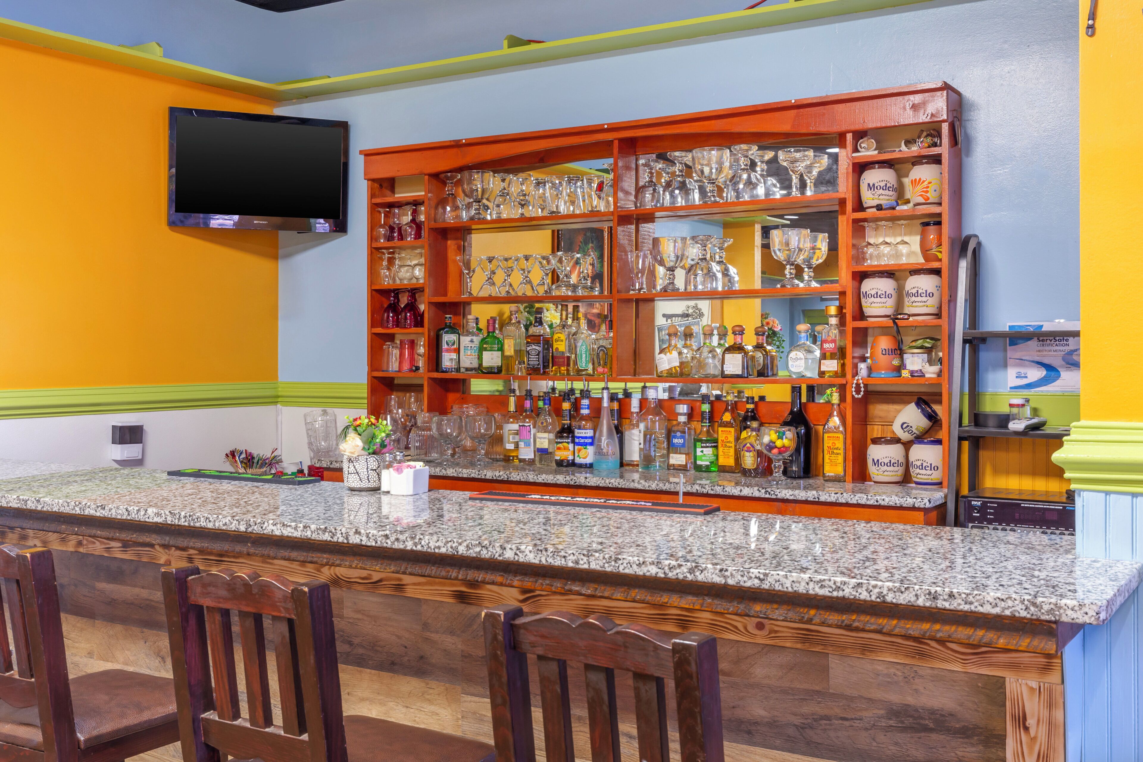 A bar area with a granite countertop, multiple liquor bottles, glasses, and a television mounted on the wall.