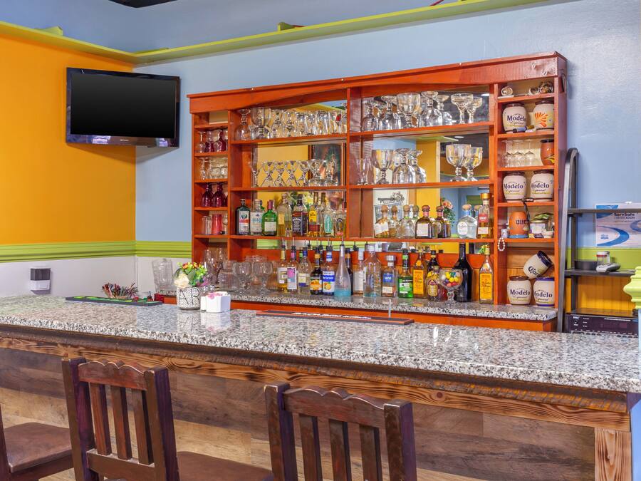 Uma área de bar com bancada de granito, diversas garrafas de bebidas, copos e uma televisão na parede.