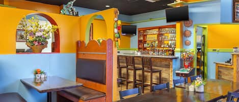A restaurant interior with wooden tables and benches, a bar area with shelves of bottles, and a television mounted on the wall.