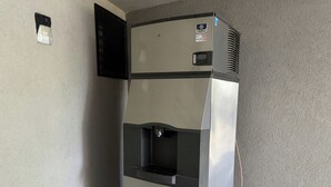 Dispensador de hielo comercial con fuente de agua, fijado a una pared de superficie texturizada.