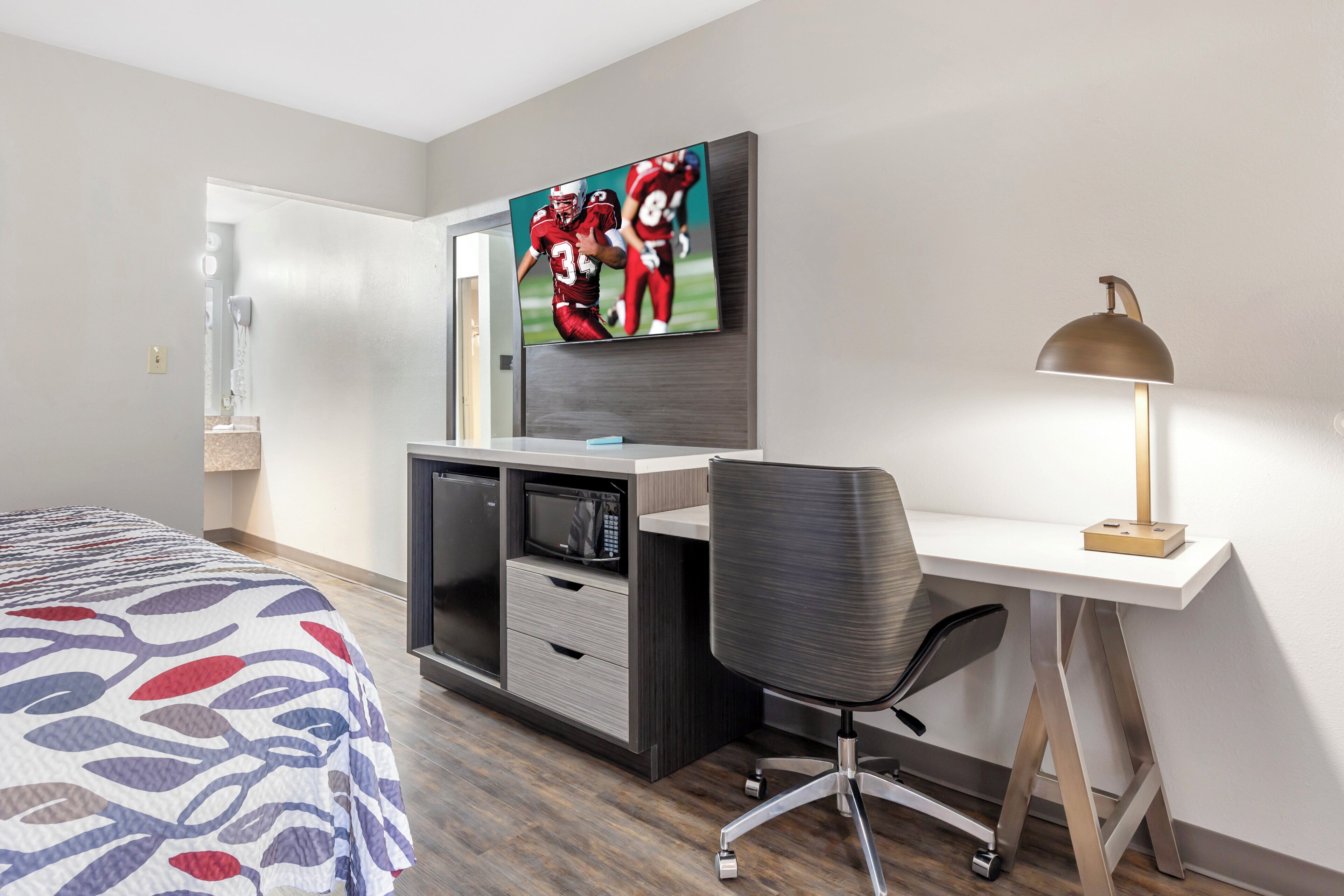A hotel room with a bed, a desk with a computer, a TV mounted on the wall, and a desk lamp.