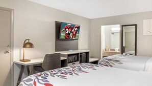 Habitación de hotel con dos camas, un escritorio con computadora, un televisor montado en la pared y un espejo.