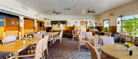 A spacious dining area with multiple wooden tables and chairs, ceiling fans, and a patterned carpet.