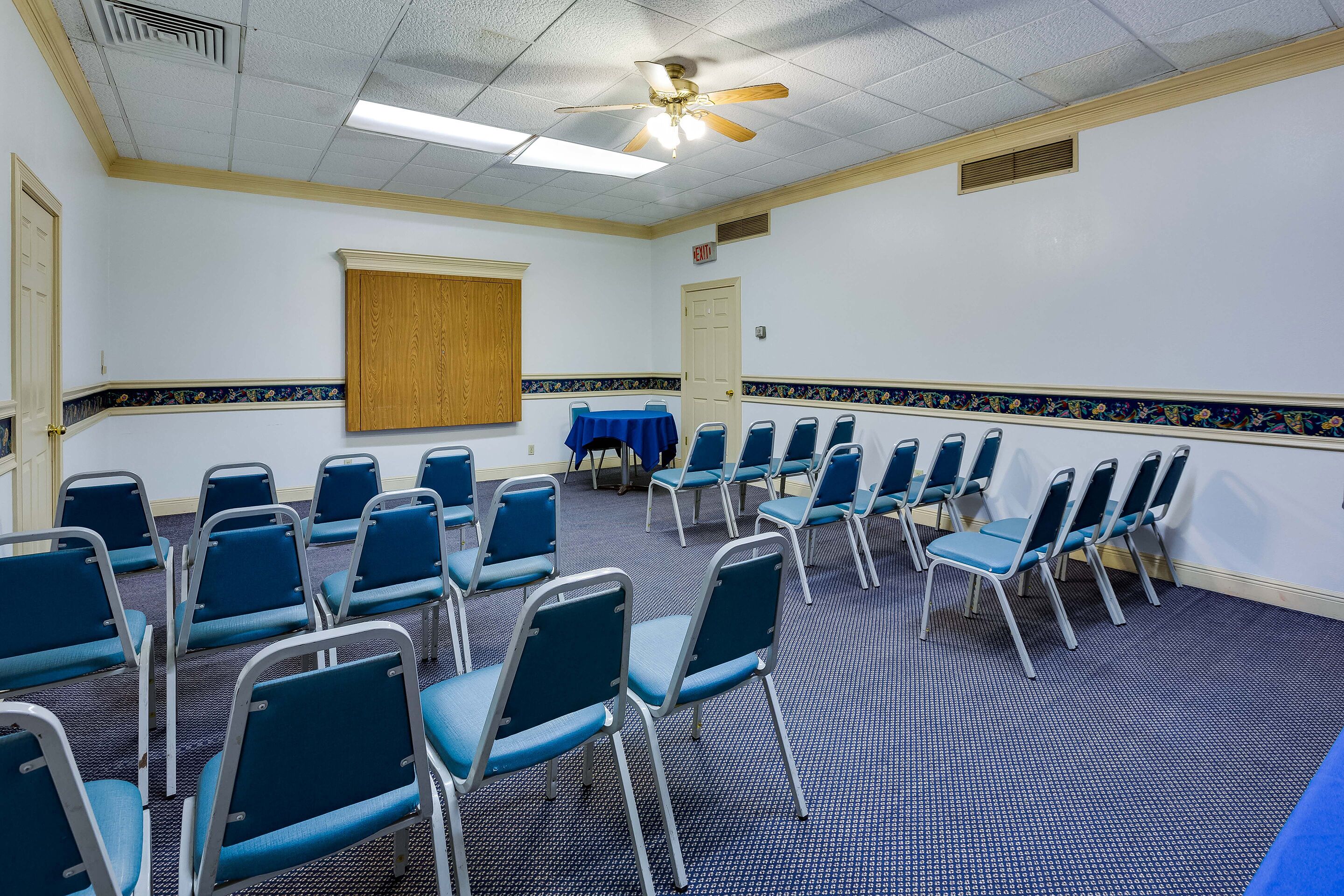 A conference room with rows of blue chairs, a ceiling fan, and a projector screen.