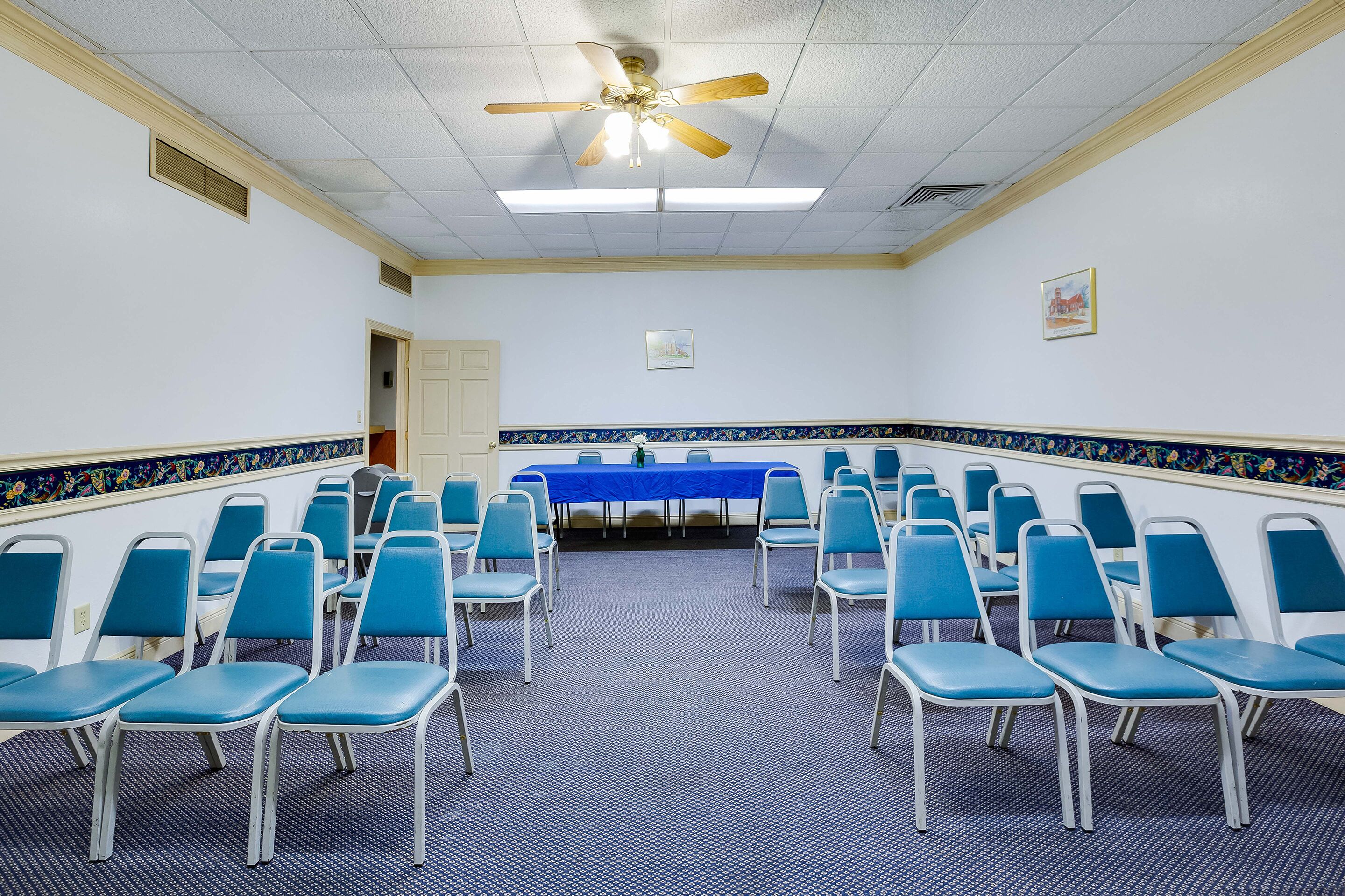 Un salón de conferencias con filas de sillas azules, una mesa azul y un ventilador de techo.