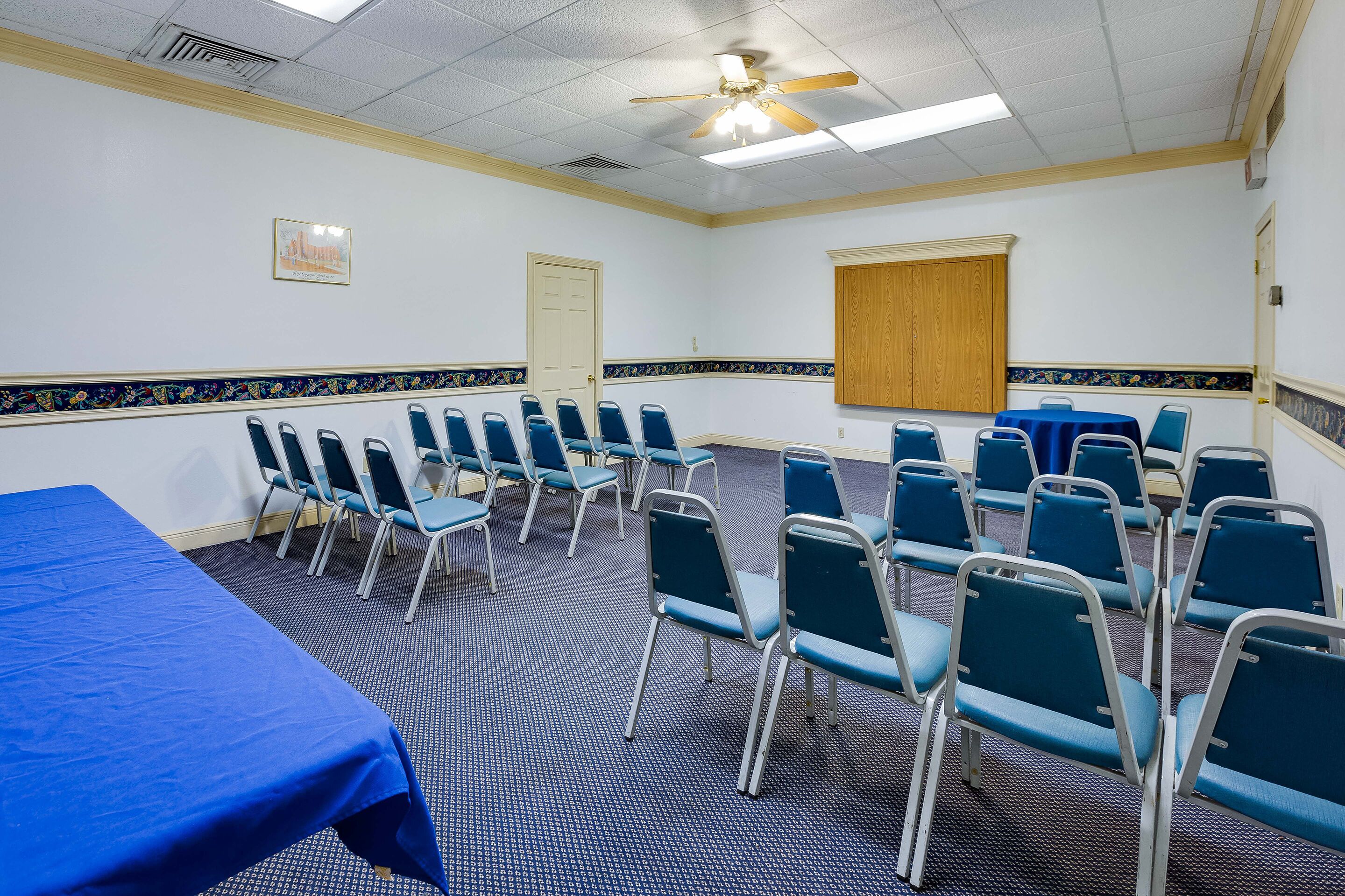 A conference room with a blue table, rows of chairs, a ceiling fan, and a projector screen.