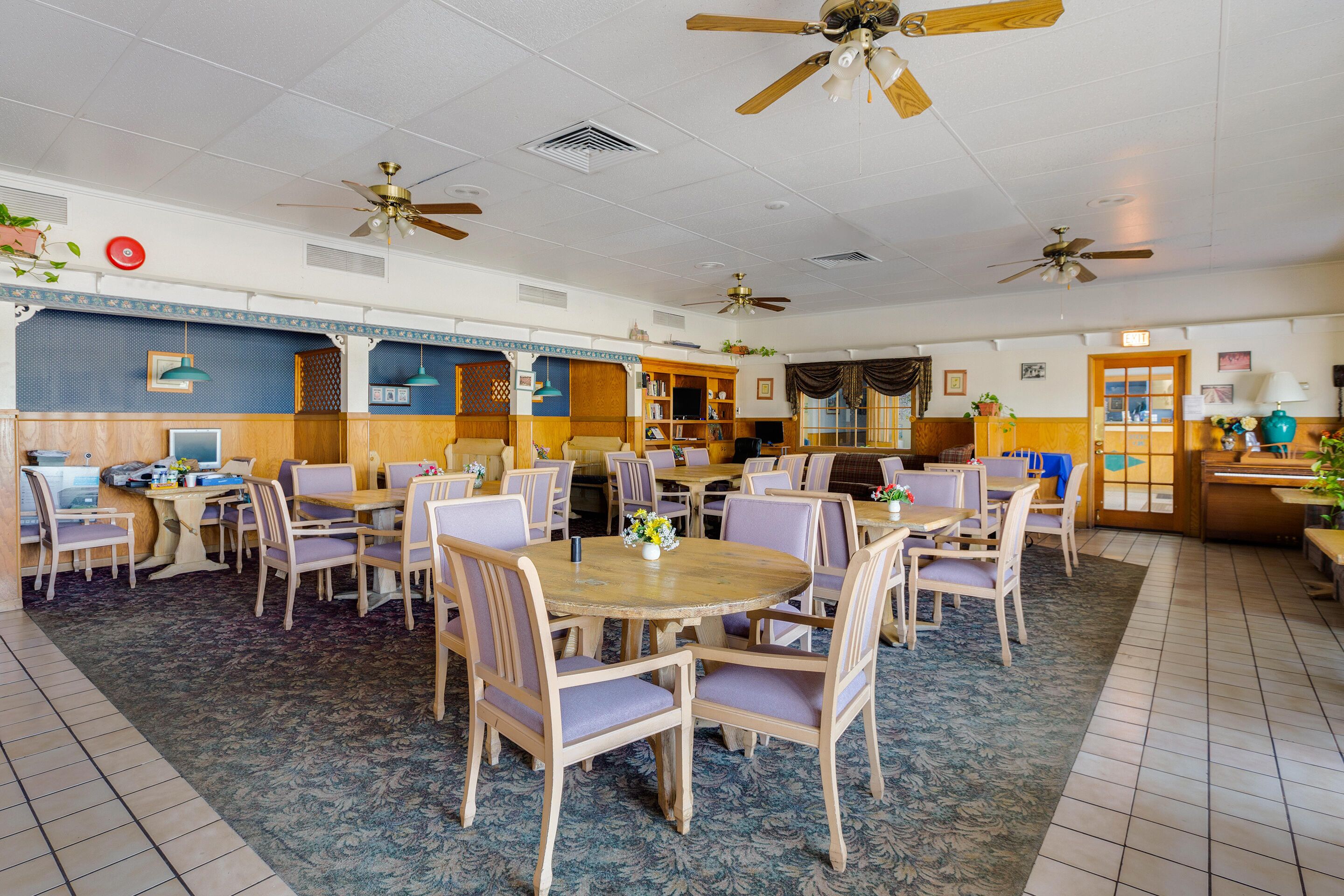 A spacious dining area with multiple tables and chairs, ceiling fans, and a kitchen area in the background.