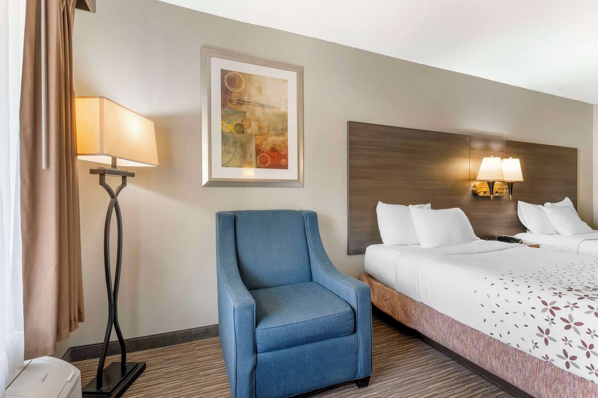 A hotel room with a bed, a blue armchair, a floor lamp, a wall-mounted lamp, and a painting.