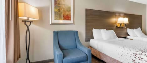 Habitación de hotel con una cama, un sillón azul, una lámpara de pie y lámparas de pared, además de un cuadro.