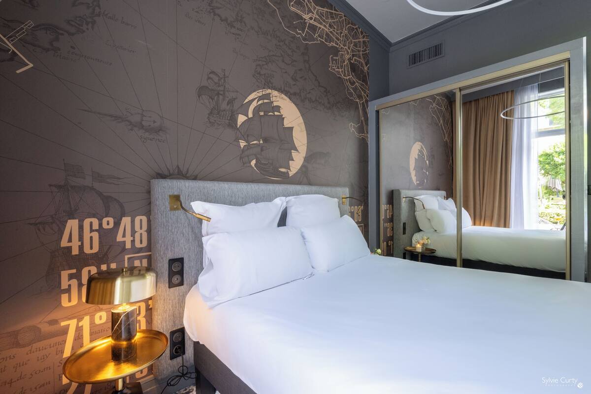 A hotel room with a large bed, a world map on the wall, a mirror, and a small table with a lamp.
