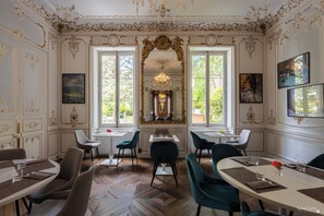 A dining area with ornate wall decorations, a chandelier, and large windows with a view of greenery.