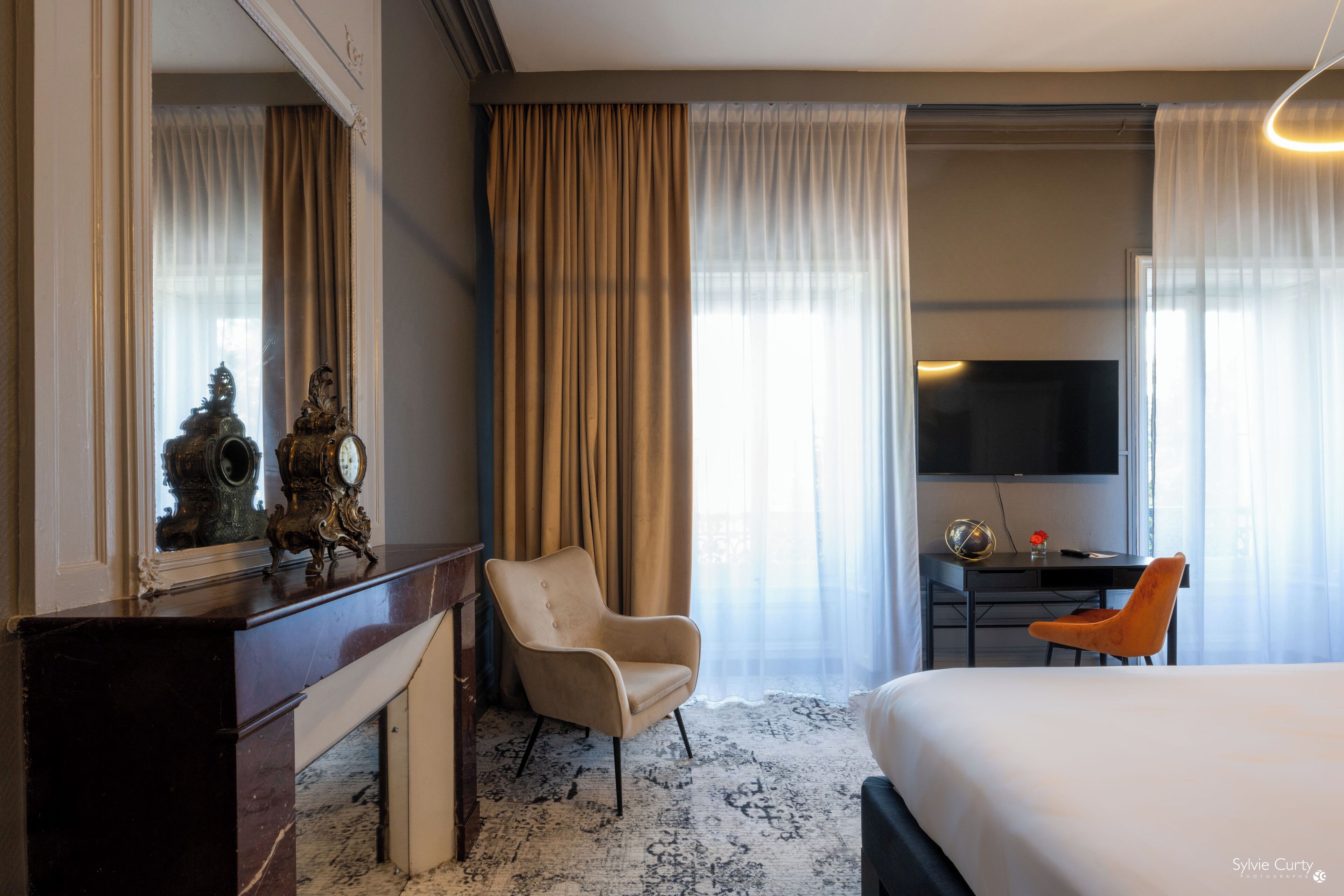 A hotel room with a bed, a desk with a TV, a chair, a dresser, and a large window with curtains.
