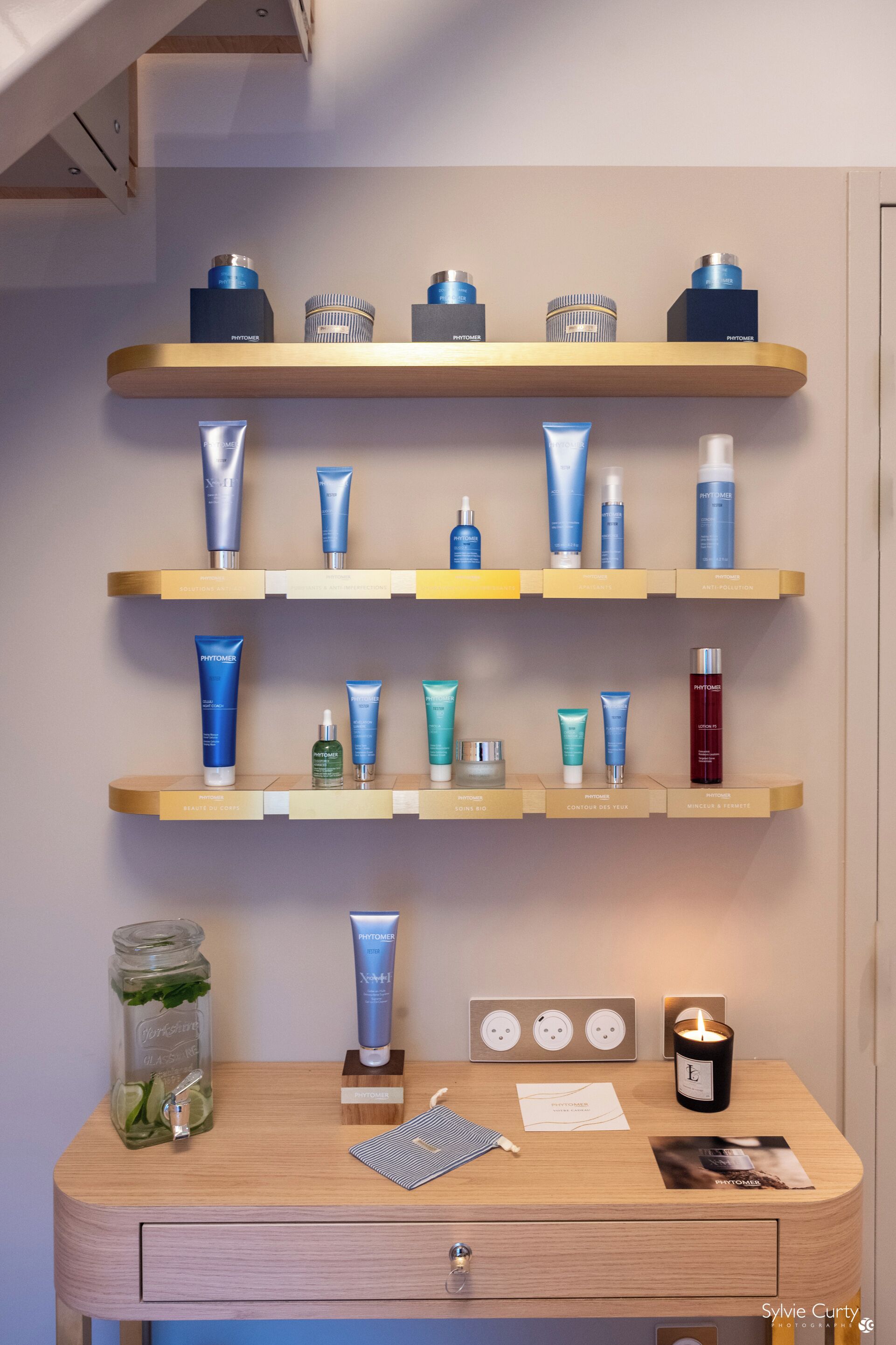 A bathroom vanity with multiple shelves displaying various skincare products, a jar of lime with mint, a candle, and a magazine.